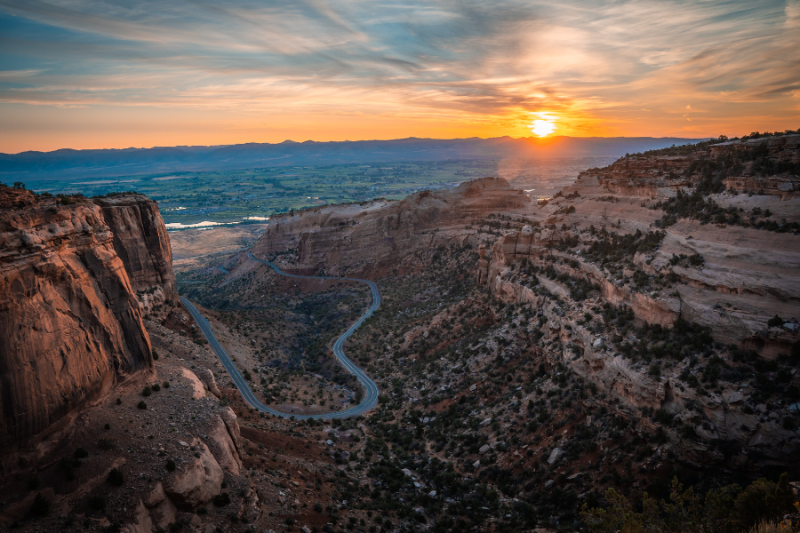 A stunning view of a winding road on the Colorado National Monument cutting through dramatic red rock canyons at sunset.
