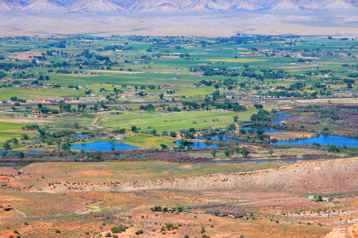 Grand Valley agricultural land with scattered trees, buildings, ponds, and reservoirs surrounded by reddish, barren land and tan mountains.