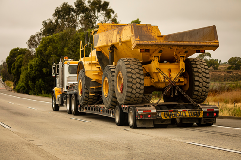 A semi-truck with a long bed is hauling an oversized construction dump truck, with a wide load banner attached to the back.