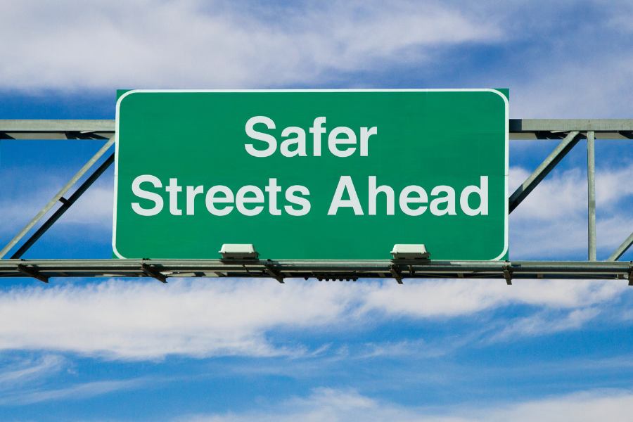 Large green street sign in front of the bright blue sky reading "Safer Streets Ahead" in white font. 