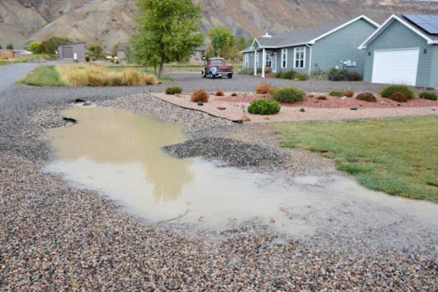 A gravel driveway and yard area with a large puddle of muddy water after rainfall, in front of a light green house with a white garage and landscaping, with hills visible in the background.