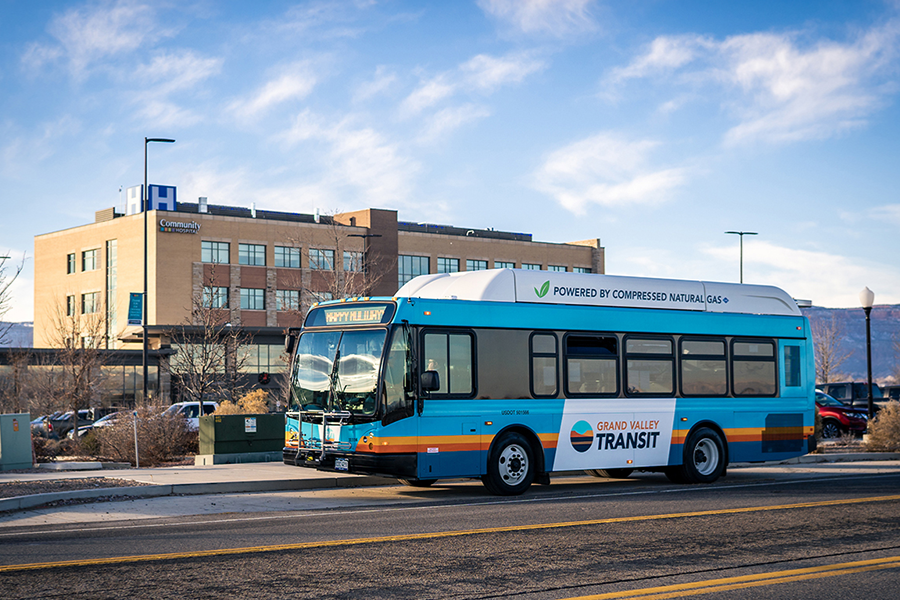 A Grand Valley Transit bus powered by compressed natural gas travels along a city street, with a medical office building and the Colorado National Monument visible in the background on a clear day.