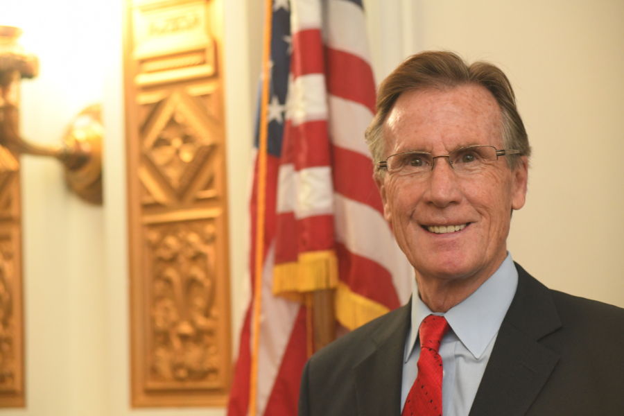 Scott McInnis stands indoors wearing a dark suit, light blue shirt and red tie, smiling toward the camera. An American flag and decorative architectural panels are visible in the background.