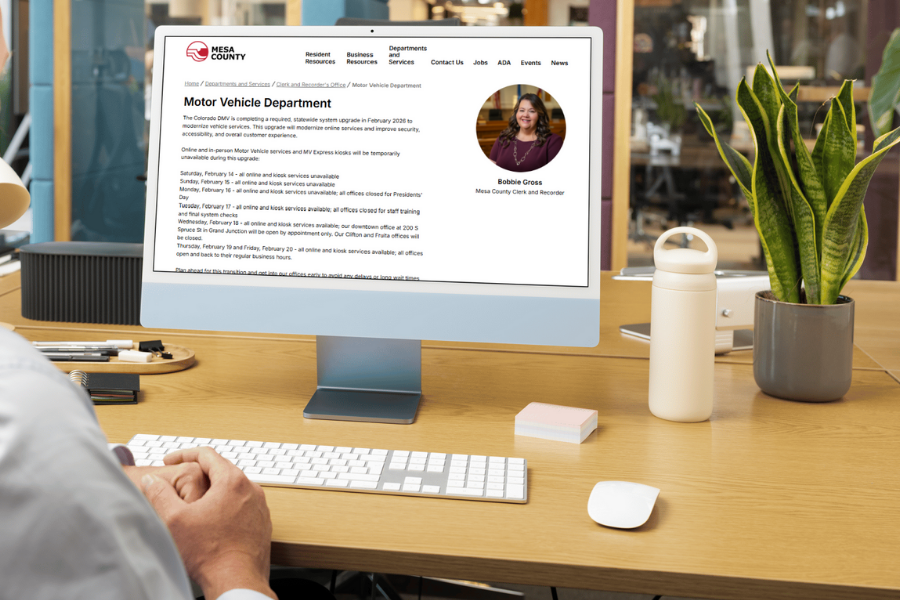 A person seated at a desk looks at a desktop computer displaying the Mesa County Clerk and Recorder website’s Motor Vehicle Department page, which outlines temporary DMV service changes during a system upgrade. The screen includes a photo of Clerk and Recorder Bobbie Gross, with office items and plants visible in the workspace.