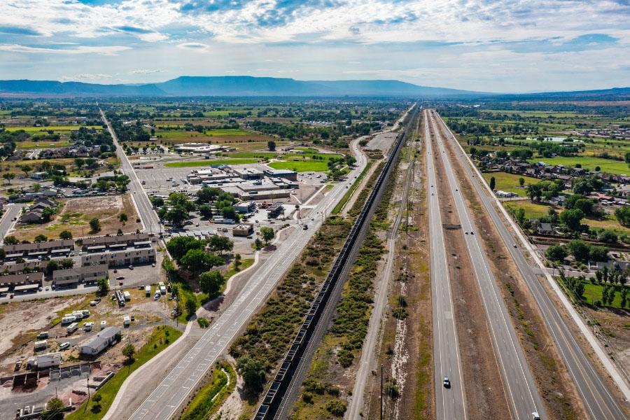 A wide aerial view of a busy transportation corridor in Mesa County, showing a multi-lane highway, a parallel road, and a railroad line running through a mix of homes, businesses, and agricultural land with mountains in the distance under a partly cloudy sky.