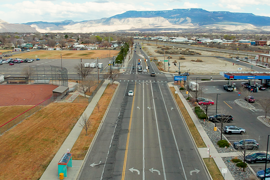 Aerial view of Orchard Avenue showing a multi-lane roadway, signalized intersection, nearby businesses and ballfields, with surrounding neighborhoods and the Grand Mesa in the background.