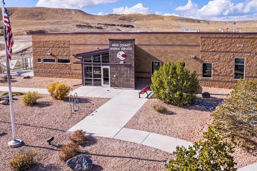 Exterior view of the Mesa County Animal Services building with the facility entrance, landscaping and an American flag visible in front of the building. Desert hills rise in the background under a partly cloudy sky.