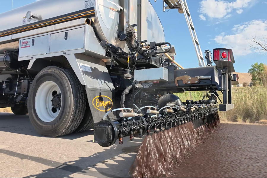 The back of a large tanker truck with paving equipment sprays sealant onto a road.