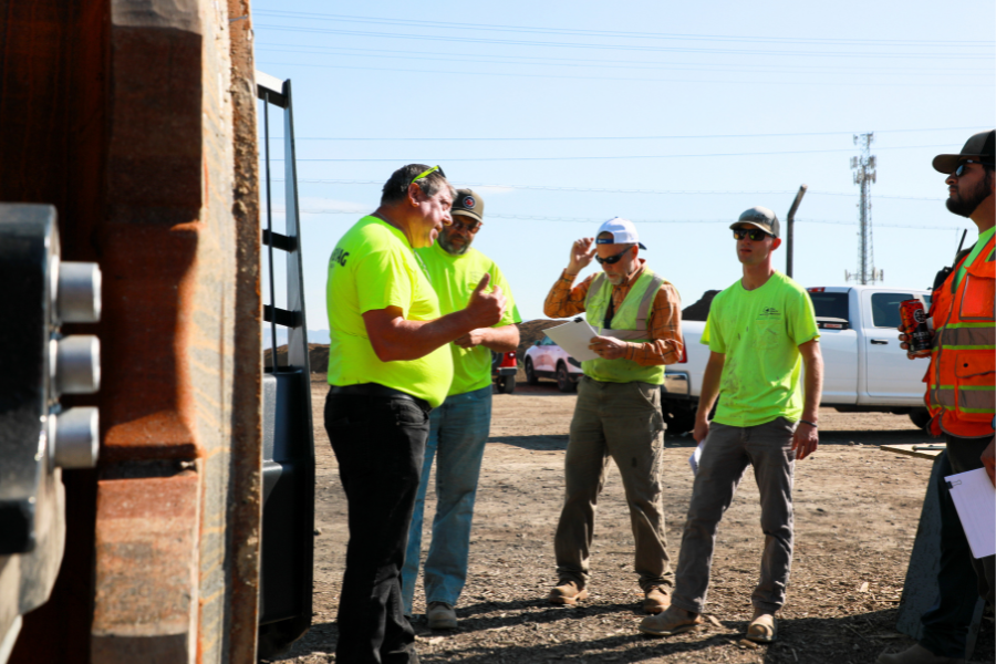 Landfill crews in high-viz shirts stand next to the wheel of a large trash compaction machine while listening to information about its operation.